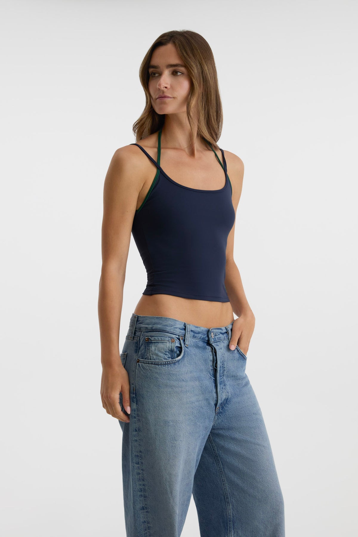 A woman modeling a navy blue tank top with thin shoulder straps and light-wash blue jeans against a plain light gray studio background.